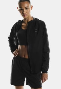 Must must lõngusjope koos kapuutsiga, esine tõmblukk ja logo detailidega, kombineeritud musta spordirinnahoidja ja lühikestega. Sile tekstuur, sportlik lõige.