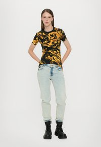 Svart t-skjorte med gul barokk mønster, korte ermer; sammen med lyseblå, litt baggy jeans og svarte snøresko.