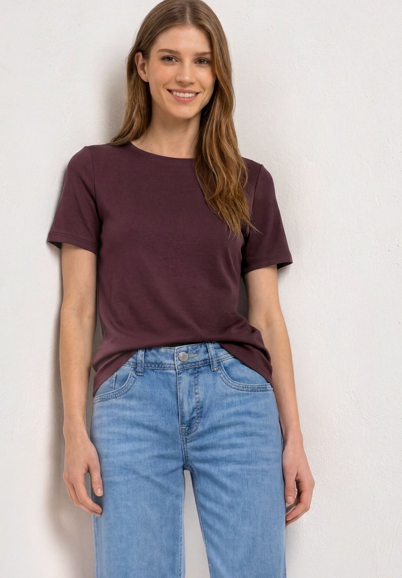 Jeune femme aux longs cheveux bruns portant un t-shirt uni violet foncé à manches courtes et un jean bleu clair, debout contre un mur blanc.
