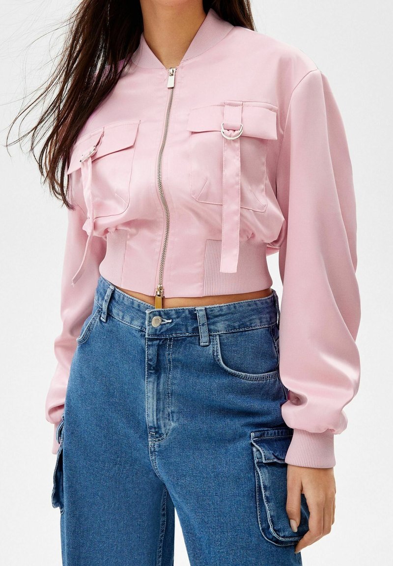 Ljust rosa kort bomberjacka med dragkedja, två framfickor och ribbade muddar, tillsammans med blå högmidjade jeans.
