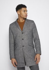 Manteau en mélange de laine gris au design ajusté, avec des revers à cran, deux poches avant et une fermeture à trois boutons. Superposé sur un pull en tricot marron.