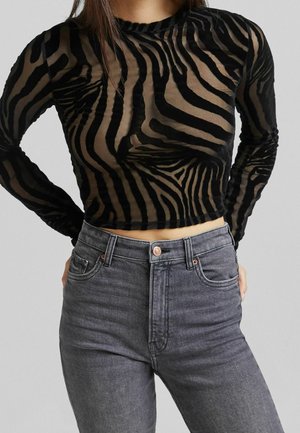 Vrouw draagt een zwart en doorzichtig zebraprint cropped lange mouwen top gecombineerd met hoog getailleerde vervaagde zwarte jeans, staand met handen in de zij.