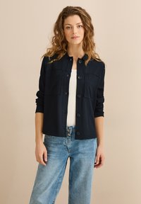 Giacca button-up blu navy con tessuto testurizzato, due tasche sul petto e colletto. Abbinata a jeans in denim azzurro chiaro a gamba larga.