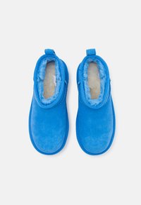 Des pantoufles bleu en suede avec un intérieur doux et moelleux, une forme arrondie, et des languettes à l'arrière pour un enfilage facile. Texture lisse sur toute la surface.