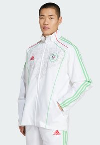 Vit sportjacka med gröna och röda accenter, prydd med ett blommönster, med dragkedjeförslutning och Algeriets fotbollslandslags emblem.