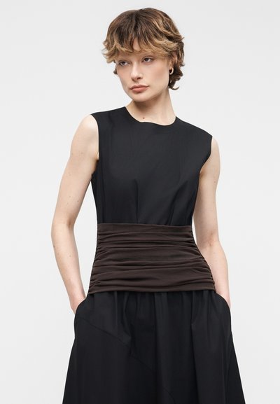 Ženska s kratkimi valovitimi lasmi, oblečena v brezrokavno črno obleko z nagubanim širokim temno rjavim pasom, roke v žepih, nevtralna ozadje.