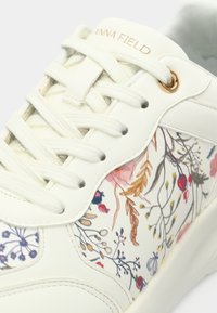 Baskets blanches avec motif floral sur le côté, lacets crème et marquage "ANNA FIELD" sur la languette en lettres dorées.