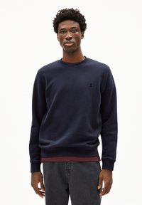 Marineblauwe sweatshirt van een katoenmix, met een ronde hals en een klein logo op de borst, gedragen met een bordeauxrood shirt eronder.