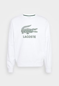 Sudadera blanca con un logo de cocodrilo verde y "LACOSTE" en texto verde a juego, con un diseño de cuello y puños de canalé.