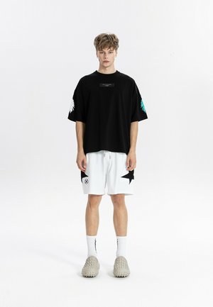 Joven con una camiseta negra holgada con estampados en las mangas, pantalones cortos blancos con detalles de estrellas negras, calcetines blancos y zapatos beige sin cordones con textura.