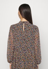 Femme aux longs cheveux foncés portant une robe à manches longues avec un motif floral, une petite ouverture en forme de trou de serrure dans le dos et un bouton à l'encolure.
