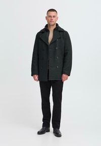 Mørkegrå uldblanding peacoat med dobbeltbrystet design, store knapper og spids krave, lagd oven på en beige sweater og sorte bukser.