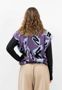 Lila und blaue gemusterte Fleece-Poncho mit weiten Armöffnungen, kombiniert mit einem langen schwarzen Oberteil und beigen Hosen, die einen lässigen Schnitt und eine weiche Textur zeigen.