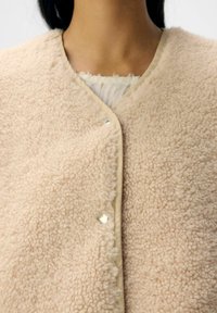 Beige fleecejack met een zachte, textuurrijke oppervlakte, voorzien van een eenvoudig ontwerp, v-hals en een drukknoopsluiting aan de voorkant.