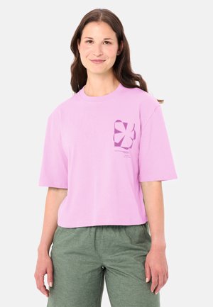 Jonge vrouw met lang bruin haar, gekleed in een losse lavendelkleurige T-shirt met een paarse print en olijfgroene broek, staand tegen een witte achtergrond.