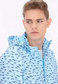 Hellblaue Jacke mit einem Dinosaurierdruck in Dunkelblau. Verfügt über einen hohen Kragen, einen Reißverschluss und eine Kapuze mit verstellbaren Kordeln.