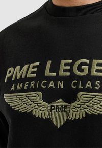 Černá mikina s výrazným olivově zeleným vyšitým textem: "PME LEGEND AMERICAN CLASSIC" s designy křídla a štítu.