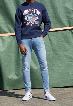 Man draagt een marineblauwe Manhattan Tennis Club-trui, lichtblauwe spijkerbroek en witte sneakers, staand met één hand in de zak.