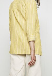 Żółta bluzka w paski o luźnym kroju, z rękawami do połowy przedramienia i jedną kieszenią, zestawiona z beżowymi spodniami o szerokich nogawkach.