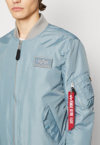 Light blue bomber jacket with a gray ribbed collar, front zip, embroidered logo patch, and a red tag reading "REMOVE BEFORE FLIGHT."