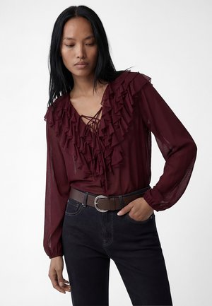 Kobieta w przezroczystej bordowej bluzce z warstwowymi falbanami i sznurowanym przodem, w czarnych dżinsach i brązowym pasku, pozująca z ręką w kieszeni.