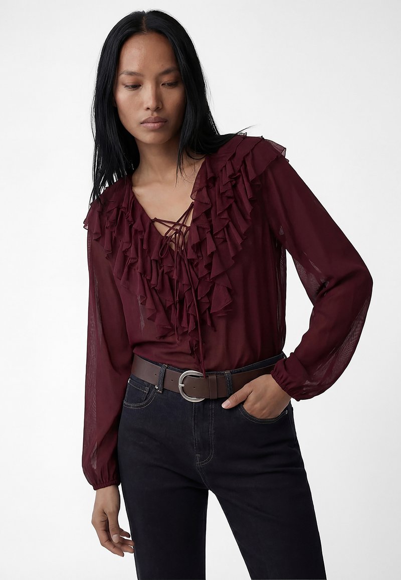 Mujer con blusa burdeos translúcida con volantes superpuestos y detalle de cordones en el frente, combinada con jeans negros y cinturón marrón, posando con la mano en el bolsillo.