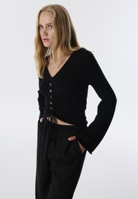 Zwarte geribbelde gebreide top met een V-hals, lange klokmouwen en een strik aan de voorkant. Heeft decoratieve letters in het midden en een cropped zoom.