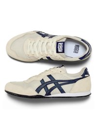 Scarpe da ginnastica beige con dettagli blu navy, caratterizzate da una tomaia in suede e tessuto, allacciatura tradizionale e suola in gomma testurizzata.