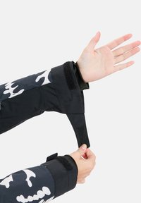 Manga de vestuário exterior em preto e branco com fecho em velcro, apresentando uma textura suave e um padrão abstrato em branco. Ajuste da alça visível à mão.