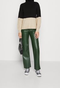 Maglione a collo alto a righe nere e beige, pantaloni ampi in ecopelle verde e borsa nera con accessori in metallo argentato. Sneakers bianche con accenti neri.