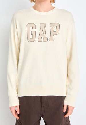 Cremefarbener Strickpullover mit einem runden Halsausschnitt. Besitzt ein großes, braunes "GAP"-Logo über der Brust. Rippbündchen und Saum.
