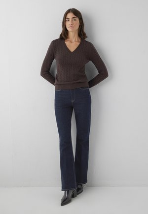 Woman standing against a wall wearing a brown V-neck cable knit sweater, dark blue bootcut jeans, and black heeled ankle boots.
