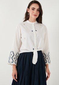 Camisa blanca de botones con bordado azul y dorado en las mangas acampanadas, atada a la cintura. Combinada con una falda plisada azul marino.