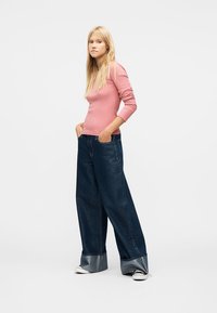 Giovane donna con lunghi capelli biondi che indossa una maglietta rosa a maniche lunghe, jeans larghi blu scuro con risvolti e sneakers bianche e nere, con le mani nelle tasche.