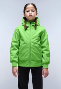 Giacca verde con cappuccio, realizzata in un tessuto liscio e resistente all'acqua, dotata di zip frontale e ampie tasche anteriori. Abbinata a pantaloni neri.