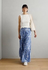 Weißes Schürzentop kombiniert mit einem blauen Maxirock mit Blumenmuster. Der Rock hat ein gestuftes Design und eine fließende Textur, ergänzt durch weiße Sneaker.