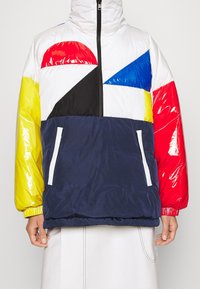 Padded jacket with high collar featuring multicolored panels: red, blue, white, yellow, black. Includes a navy pocket with white zipper.