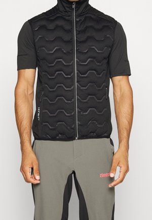 Mann trägt eine schwarze gesteppte ärmellose Weste über einem schwarzen kurzärmeligen Hemd und graue Hosen mit pinkem Logo am Oberschenkel.