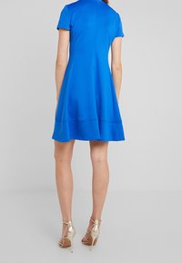 Kortärmad, utsvängd blå klänning med en slät yta och en dold dragkedja bak; ihop med metalliska högklackade sandaler.