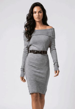 Mujer con cabello largo y oscuro, vistiendo un vestido gris ajustado sin hombros, con mangas largas y un cinturón negro, de pie frente a un fondo liso.