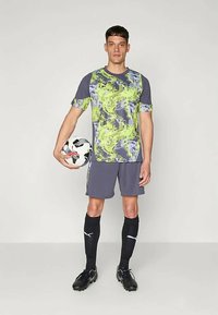 Κοντομάνικη φανέλα σε γκρι χρώμα με πράσινα αφηρημένα σχέδια, συνδυασμένη με γκρι σορτς, μαύρες κάλτσες μέχρι το γόνατο και μαύρα παπούτσια ποδοσφαίρου, κρατώντας μια μπάλα ποδοσφαίρου.