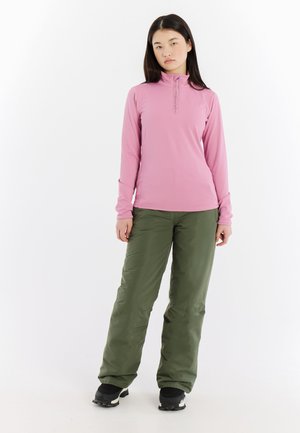 Langärmliges, rosa Sportoberteil mit einem halben Reißverschluss am Kragen, kombiniert mit weiten grünen Outdoor-Hosen und schwarzen Schneestiefeln.