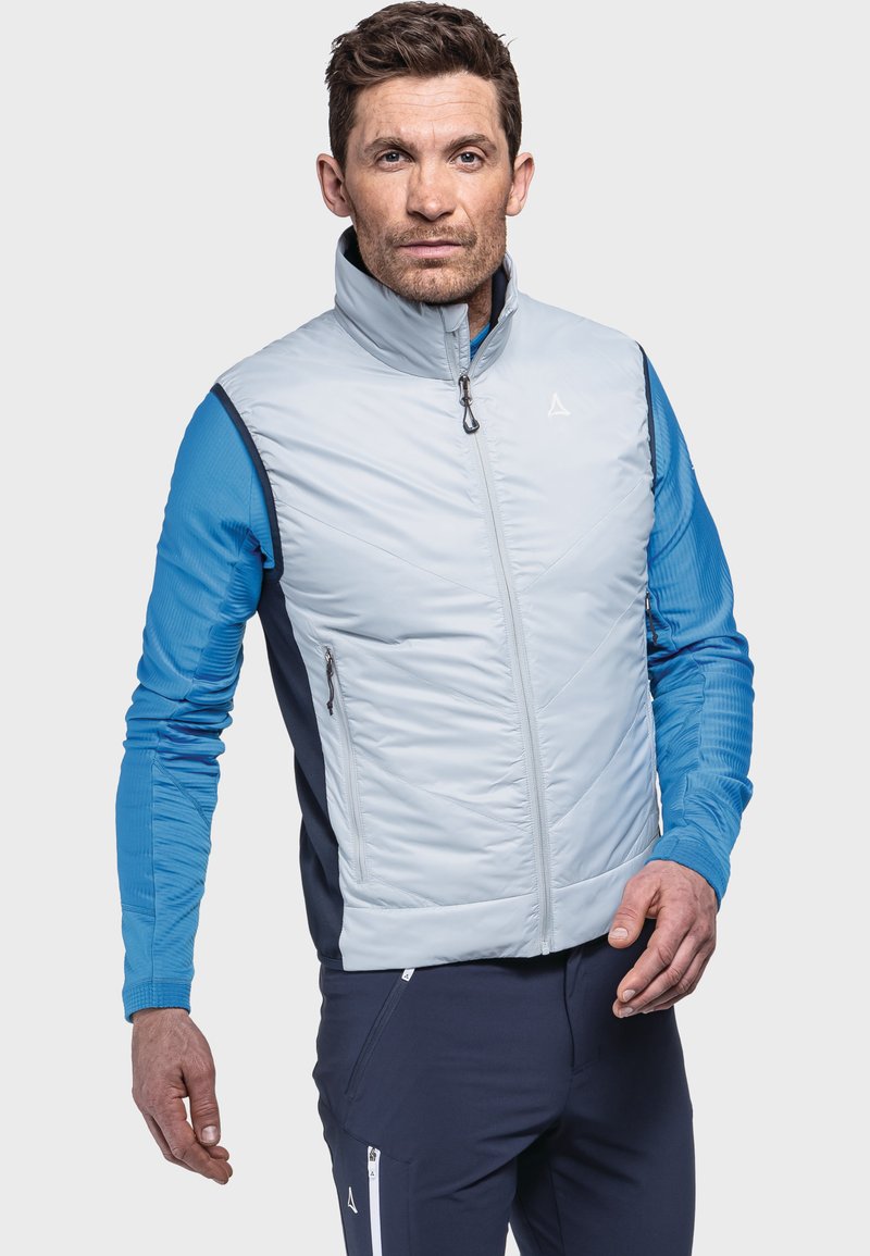 Hellgraue isolierte Weste mit hohem Kragen und Reißverschluss auf der Vorderseite, kombiniert mit einem blauen Langarmshirt und marineblauen Hosen mit Reißverschlusstaschen.