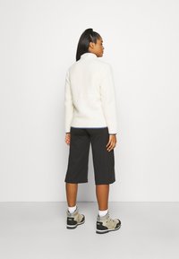 Fleece-takki off-white-värissä, jossa on korkea kaulus ja kontrastiväriset tikkaukset. Yhdistetty mustiin polvipituisiin shortsihin ja vaaleanruskeisiin vaelluskengiin.