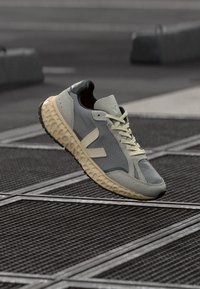 Hallide ja beežide toonidega spordijalanõud tekstuurse tallaga, mis hõljub metallvõrgupinna kohal linnalises väliruumis.