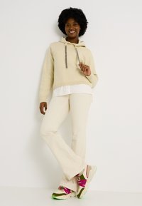 Sudadera beige con cordones, superpuesta sobre una camisa blanca. Pantalones acampanados en beige claro. Zapatillas gruesas en verde, rosa y detalles en beige.