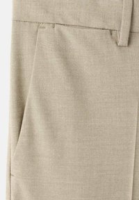 Tela de color beige claro con una textura tejida, que incluye un bolsillo lateral y detalles de costura en la cintura para un ajuste ceñido.