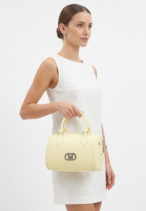 Frau in weißem ärmellosem Kleid hält eine hellgelbe Handtasche mit Metall-"M"-Logo und doppelten Griffen vor einfarbigem Hintergrund.
