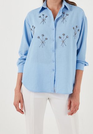 Femme portant un chemisier bleu clair boutonné avec une petite broderie florale en perles et un pantalon blanc sur un fond uni.