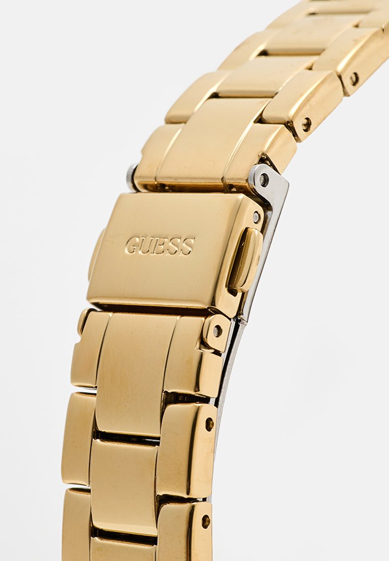 Montre-bracelet en métal doré avec un design structuré et en maillons. Dotée d'un fermoir avec un accent argenté et du logo "GUESS" gravé.